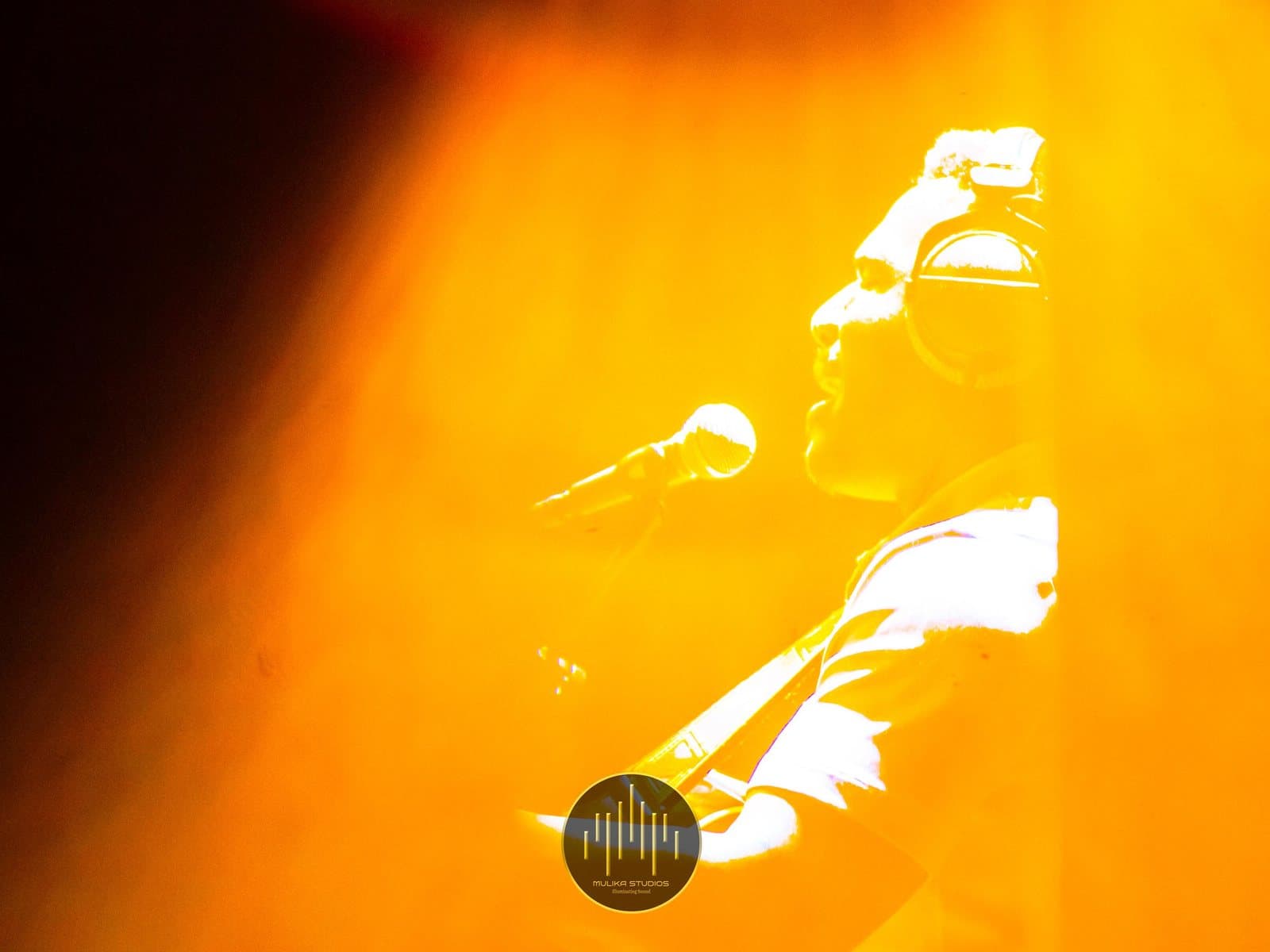 A performer singing and playing guitar under amber stage light during the Tales of Mugabi live recording